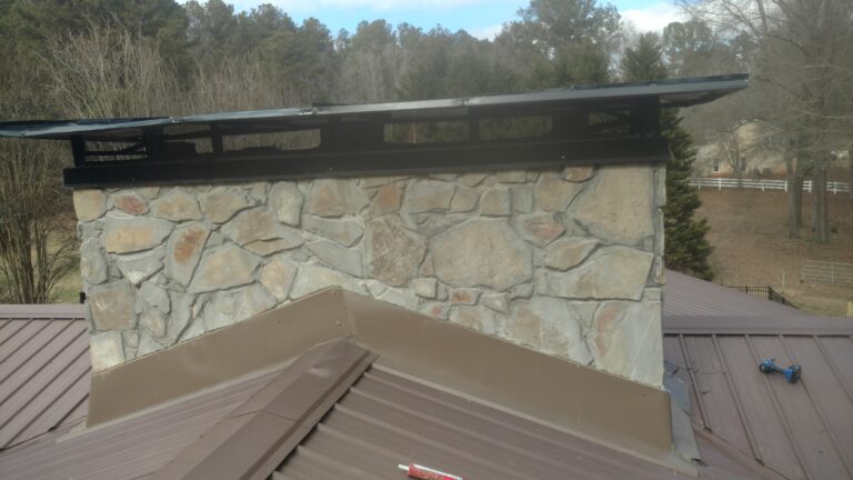 Close-up of a stone chimney with a newly installed black chimney cap and brown metal roofing, completed by Thaxton Construction in Griffin, GA. Power tools visible on the roof