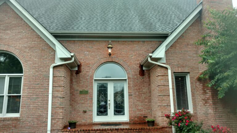 Front view of a brick home with a steep asphalt shingle roof, white double doors with decorative glass, and newly installed white gutters and downspouts by Thaxton Construction in Griffin, GA