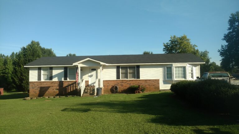 White single-story house with a dark roof and brick foundation, featuring a small front porch and a grassy lawn
