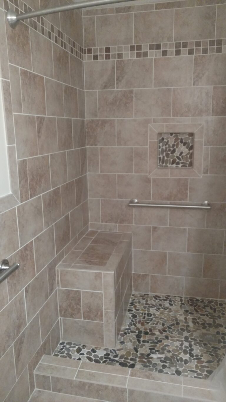 A tiled shower featuring light brown rectangular tiles on the walls and a mosaic of smooth, dark pebbles on the floor.