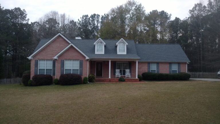 Front view of a brick house with a dark gray Asphalt Shingles roof, multiple dormer windows, and a covered front porch with white rocking chairs.