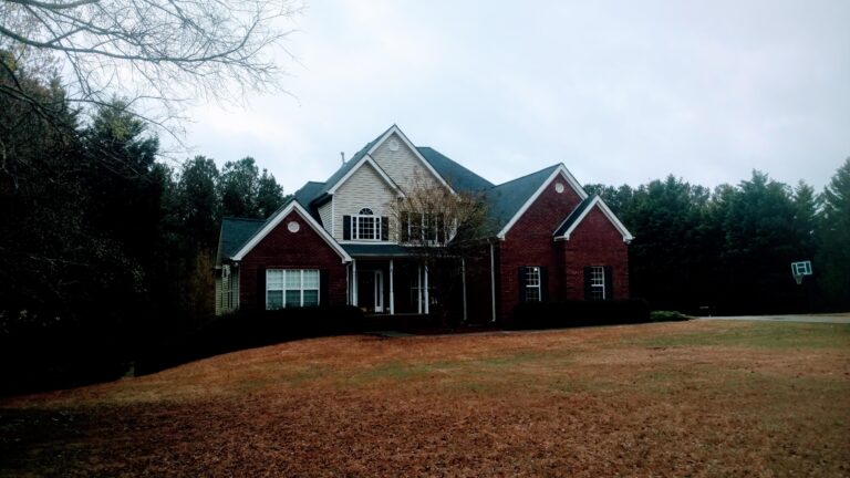 A large, two-story brick house with a dark roof and light-colored siding on the upper level, surrounded by trees and a brown lawn, potentially requiring roofing repair in Georgia.