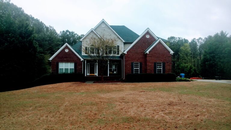Closeup view of a large, two-story brick house with a dark roof and light-colored siding on the upper level, surrounded by trees and a brown lawn, potentially requiring roofing repair in Georgia.