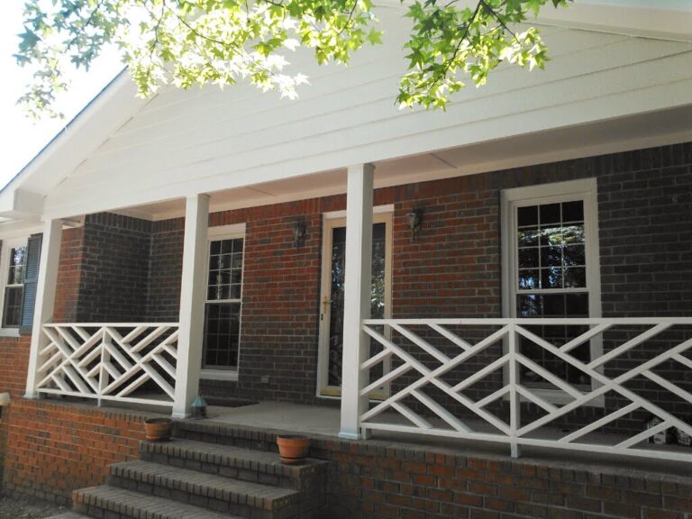A front view of a brick residential house with a white porch and decorative white railing, featuring a visible portion of the residential roofing and green foliage overhead.