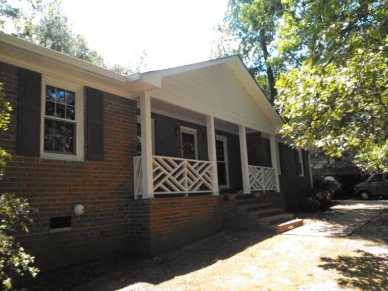 residential brick house with a white porch featuring decorative white railings and a prominent residential roofing system, surrounded by trees and a driveway