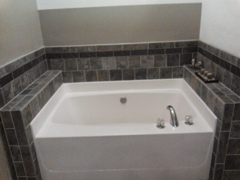 modern bathroom featuring a white built-in bathtub with chrome fixtures, surrounded by dark grey tile wainscoting and wall accents, potentially part of a renovation project by Thaxton Roofing
