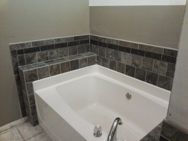 side view modern bathroom featuring a white built-in bathtub with chrome fixtures, surrounded by dark grey tile wainscoting and wall accents, potentially part of a renovation project by Thaxton Roofing