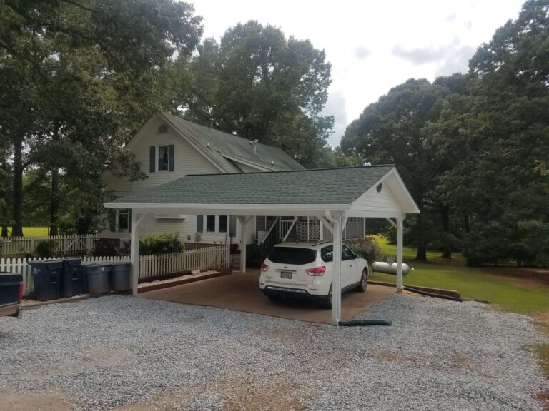 A white house with a green asphalt shingle roof and a matching green-roofed carpark covering